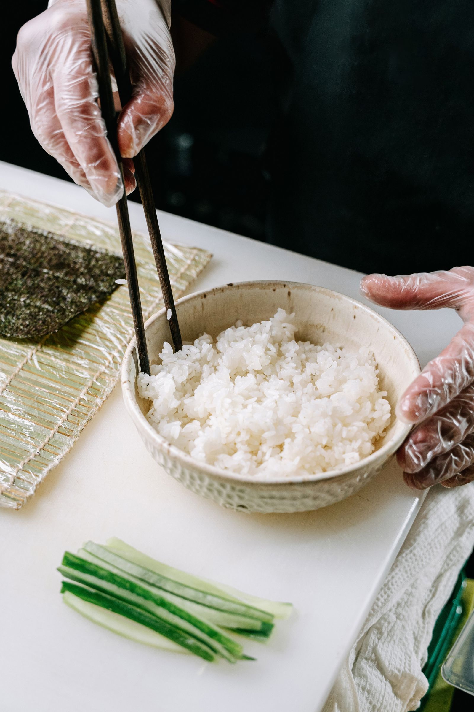 Hands preparing sushi rice beside nori and cucumber